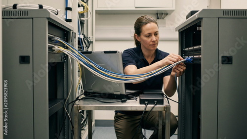 A small enterprise IT administrator connects patch cables between two compact server racks beside a laptop and external backup drives, showing hybrid cloud resilience in a practical server closet