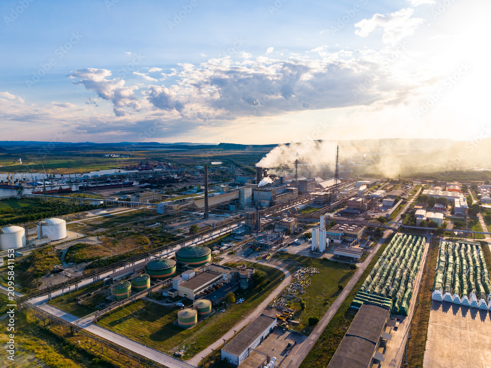 Fototapeta premium Factory complex located near a river with smoke rising from chimneys during sunset in an industrial area