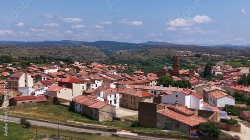 Panoramic view of La Iglesuela del Cid, a medieval village in Maestrazgo, Teruel, Spain, featuring terracotta rooftops, stone church bell tower, and wind turbines on distant ridge