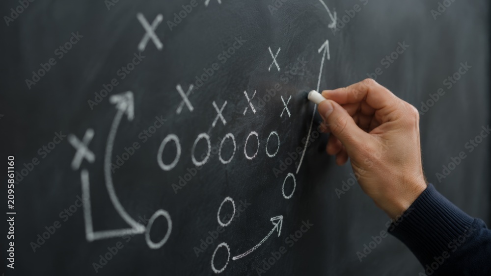 Obraz premium A close-up view of a hand drawing a football strategy diagram on a blackboard with white chalk, illustrating a coach or trainer planning a game or practice session.