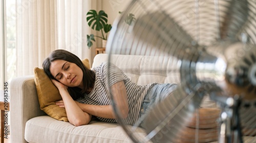 Young woman sleeping on sofa with electric fan blowing air in living room during hot weather.
