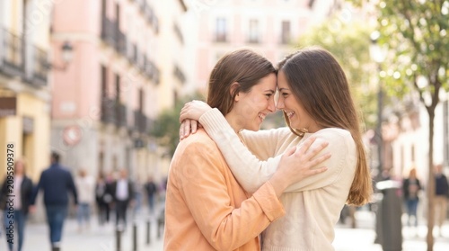Two women embrace and smile at each other on a sunny urban street. The image shows a warm, affectionate moment between friends or partners in a casual outdoor setting.