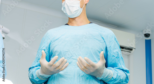 Surgeon wearing sterile gloves and blue gown holding hands up in operating room.