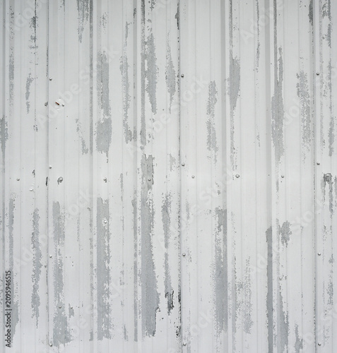 Vertical close up of a weathered white painted corrugated metal wall with peeling flaking worn paint revealing bare metal, haphazard screw fasteners, and a red screw sticking out