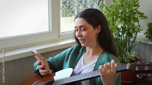 Portrait of a happy beautiful young Caucasian woman learning to play the guitar with online lessons on a smartphone. Modern remote education, digital learning, and self development concept.