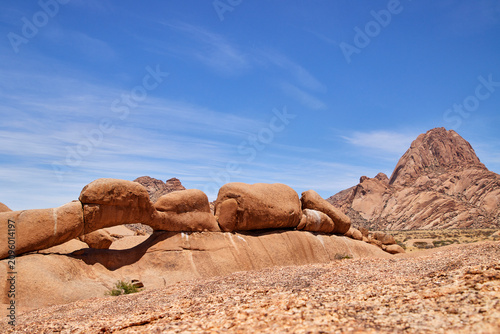 Foto Ein natürlicher Granitbogen liegt vor einer markanten Berglandschaft unter blauem Himmel an der Spitzkoppe in Namibia