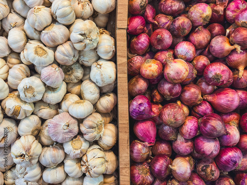 Fresh garlic heads rest beside vibrant red onions. Plump, purple-skinned shallots glow beside pearly bulbs. Market stall display invites cooks and shoppers alike