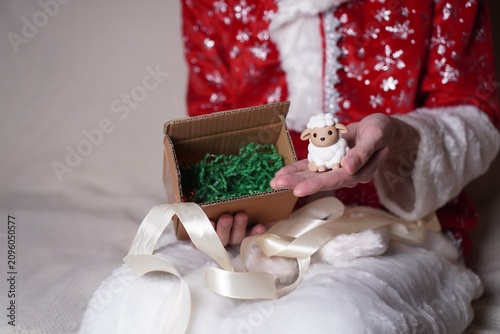 a man in a Santa Claus costume holding a box with an unpacked gift in his hands, and in another a vintage figure of a white sheep symbol of 2027 on the background of a white sofa as a New Year card 