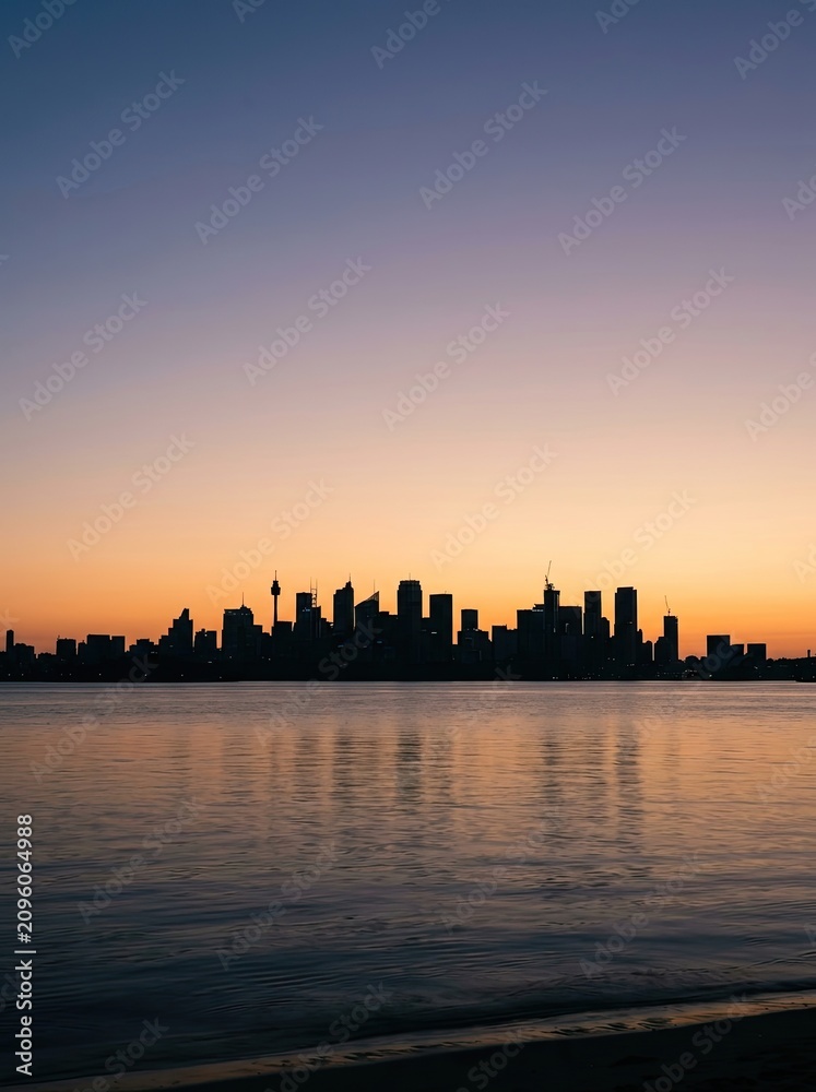 Fototapeta premium The image captures a stunning silhouette of a city skyline against a vibrant, colorful sky. A calm body of water reflects the buildings and the sunset, creating a mirrored effect.