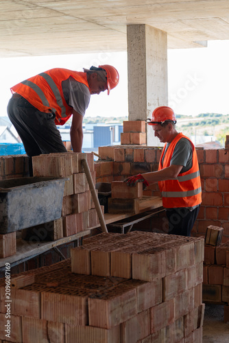 Construction worker stands on dusty ground and hands brick to colleague. Builders organize bricks neatly on construction site. Vertical shot