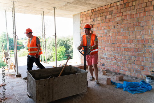 Man construction worker carefully pushes heavy cement container using sturdy wheelbarrow. Experienced builders work together efficiently