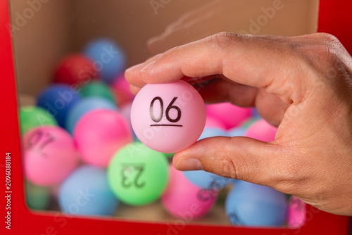 Gros plan sur une main tenant une boule de tirage au sort pour le loto, le numéro 6. En arrière-plan, une boite transparente avec des autres boules.