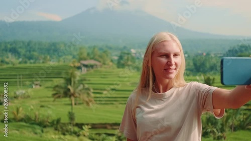 A young blonde woman smiling broadly takes a selfie with her phone in front of a stunning volcano and lush rice terraces during daytime.