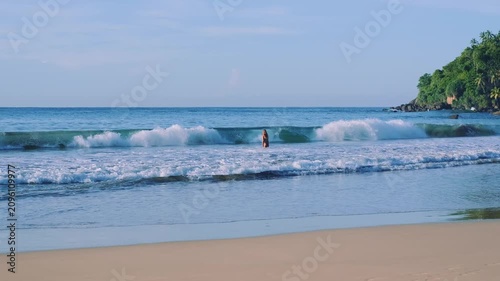 A girl carefully walks into the sea as strong waves crash near the shore at Mirissa beach on Sri Lanka island, surrounded by lush green nature and tropical scenery.