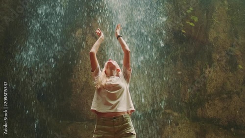 A young woman smiles joyfully with raised hands as she embraces a powerful waterfall, standing in front of the cascading water during daytime.