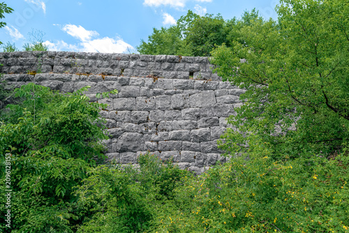 Old stone wall texture surrounded by lush green foliage in Cetinje. Ancient masonry backdrop with wild bushes and summer vegetation in Montenegro, Europe