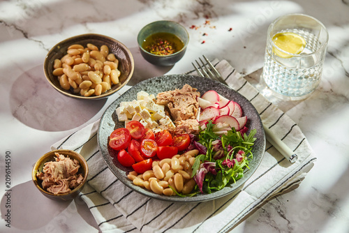 Tuna salad bowl with cannellini beans, feta cheese, cherry tomatoes, radish, and greens served on a marble table with olive oil dressing.