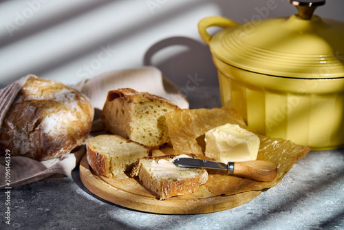 Close-up of a knife spreading butter on a slice of fresh artisan bread. Wooden board, linen cloth, and yellow pot. Homemade breakfast concept.
