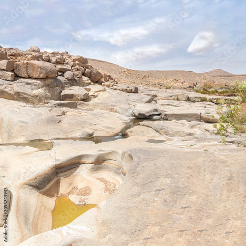 A rocky desert landscape in Afellal, Tamanrasset, Algeria, featuring a distinctive circular stone pothole filled with yellowish water and a shallow flowing stream. Blurred visitors are visible afar.