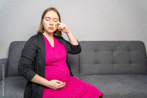 Pregnant woman sitting on a sofa while wiping away tears, expressing emotional stress, sadness, or pregnancy mood swings. Concept of prenatal mental health, emotional well-being, hormones 