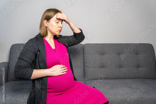 Pregnant woman suffering from headache, dizziness, or pregnancy discomfort while sitting on a sofa at home. Expectant mother touching her forehead with eyes closed, expressing fatigue, stress