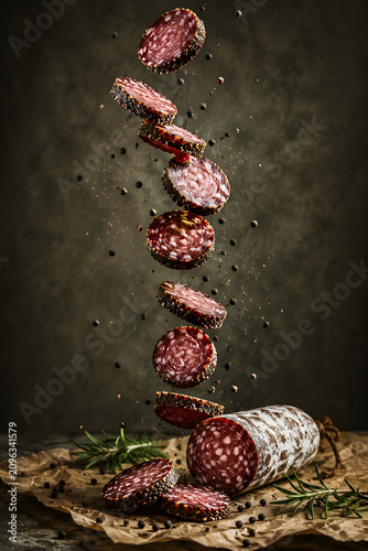 Peppered salami slices falling above whole salami with rosemary on paper