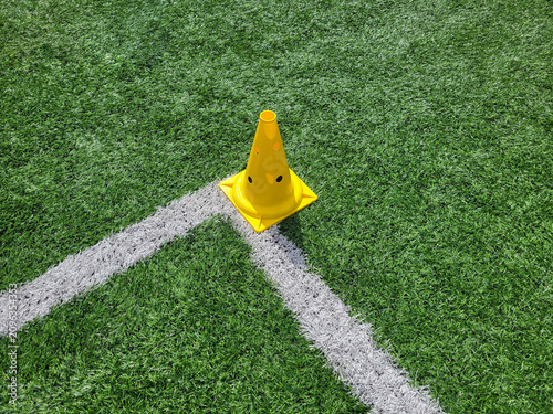 A yellow football cone stands alone on a green field near a white corner.