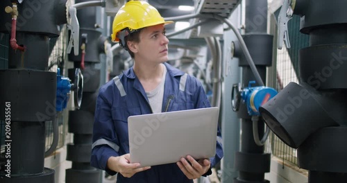 A focused industrial engineer uses a laptop to perform system diagnostics, inspecting the complex pipeline infrastructure for routine maintenance and quality control.