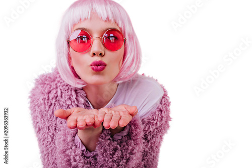 Close-up portrait of amazing female model in pink wig sending air kiss. Studio photo of good-humoured girl in sunglasses and peruke.