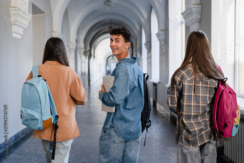 Back view of high school students walking down school corridor, teenage classmates going to class, one boy turning back and looking at camera, education and student life concept