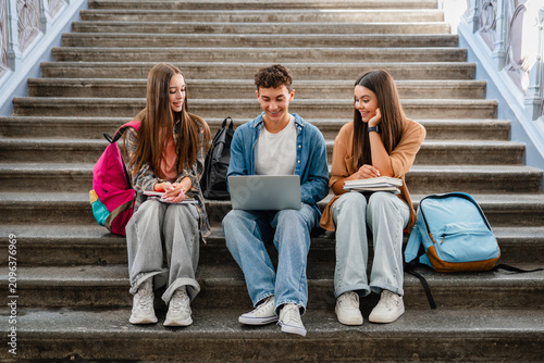 Group of high school students sitting on campus stairs with textbooks and laptop, classmates talking together, digital learning, education and student life concept