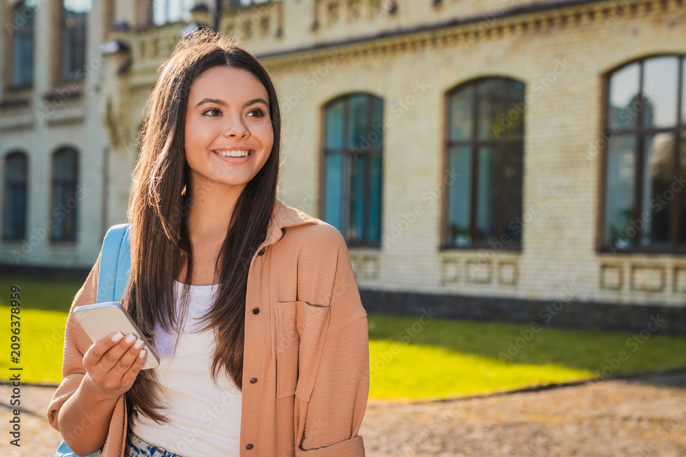 Fototapeta premium Close up of female student holding smartphone and looking at copy space in front of university building, mobile internet, online learning, digital communication and student life concept