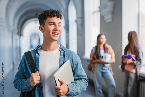 Portrait of male student with laptop in school corridor, classmates talking in background, modern education, digital learning and student life concept