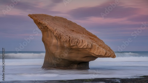 A dramatic weathered rock formation with textured layers is partially submerged in the ocean water at sunset