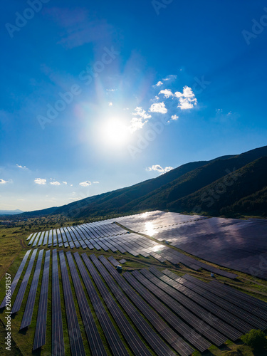 Solar panels cover a large field under bright sunlight with a blue sky and mountain backdrop in the afternoon