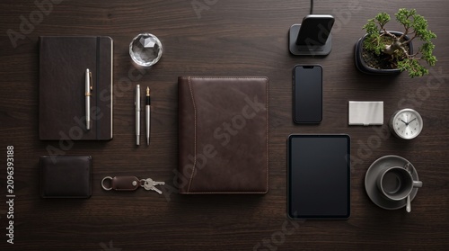 Top down flat lay view of wooden office desk with brown leather notebook pen smartphone wireless charger and small bonsai tree decoration