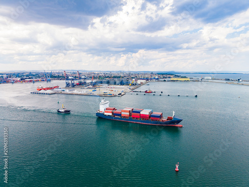 Bild auf Leinwand Large cargo ship sails through the waters near a busy port while a tugboat assis