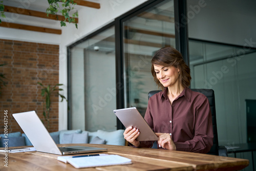 Working on digital tablet in modern office, latin mature businesswoman reads report online. Focused woman startup founder using device at desk. Female owner grows business company at workplace.
