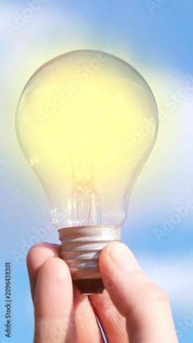Close-up of a flickering glowing light bulb held against blue sky. Symbol of ideas, innovation, unstable electricity, inspiration, creativity and renewable energy