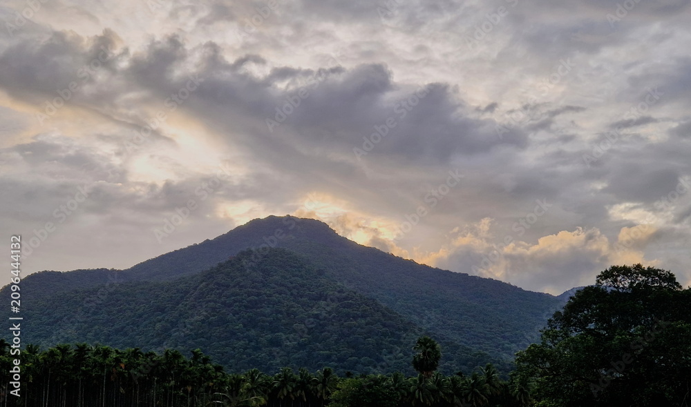 Obraz premium The sacred Velliangiri mountain peak covered in lush green forest under a dramatic stormy sky with golden sun rays breaking through heavy clouds