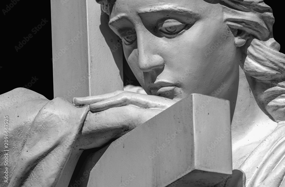 Obraz premium Black and white close-up of a mourning cemetery angel resting on a stone cross. A timeless symbol of grief, remembrance, faith, hope, eternal love and divine comfort.