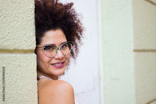 Εκτύπωση καμβά Joven mujer latina con gafas asomándose por una pared y sonriendo a la cámara, gran espacio para texto