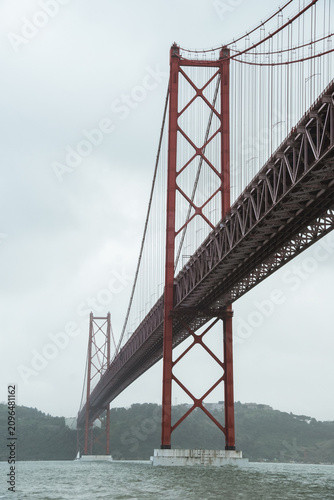 View of the 25 de Abril Bridge in Lisbon stretching across the Tagus River on a cloudy day, with soft mist surrounding the iconic red suspension structure and calm urban waterfront scenery.