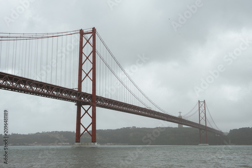 Close view of Lisbon’s 25 de Abril Bridge spanning the Tagus River, with the Cristo Rei monument visible through misty weather beneath a gray overcast sky.