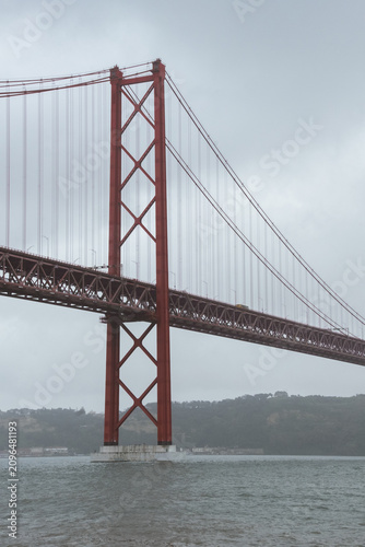 View of the 25 de Abril Bridge in Lisbon stretching across the Tagus River on a cloudy day, with soft mist surrounding the iconic red suspension structure and calm urban waterfront scenery.