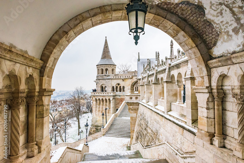 Quadro su tela Beautiful winter view of Fisherman's Bastion at Buda Castle in Budapest, framed by a classic stone archway with a hanging lantern and snow-covered stairs
