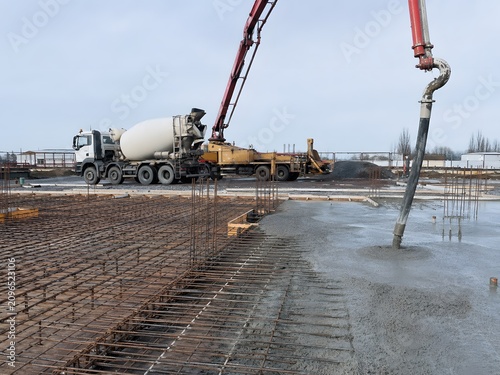 Concrete truck and pump pouring cement on construction site. Industrial machine placing fluid material onto steel rebar reinforcement mesh for building foundation. Civil engineering project.