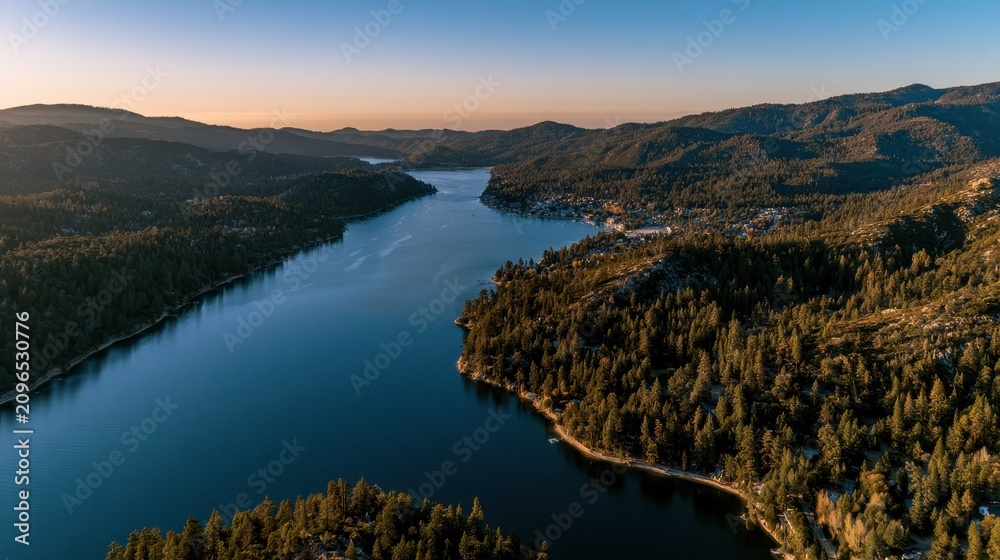 Obraz premium Golden hour drone panorama over a scenic alpine lake with forested shores and a distant lakeside community