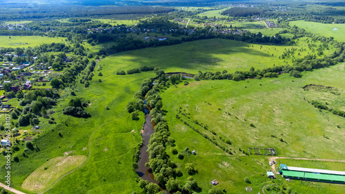 Lush green fields stretch wide under a bright sky. Narrow river meanders through the landscape. Distant villages feature clustered homes and trees. Green tent sits near the riverbank