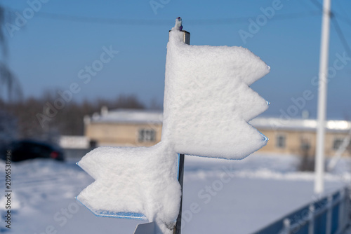 Snow covers a traffic light arrow shape. Winter's quiet stillness surrounds the scene. Snow blankets the sign and distant houses. Clear blue sky contrasts with the wintry landscape